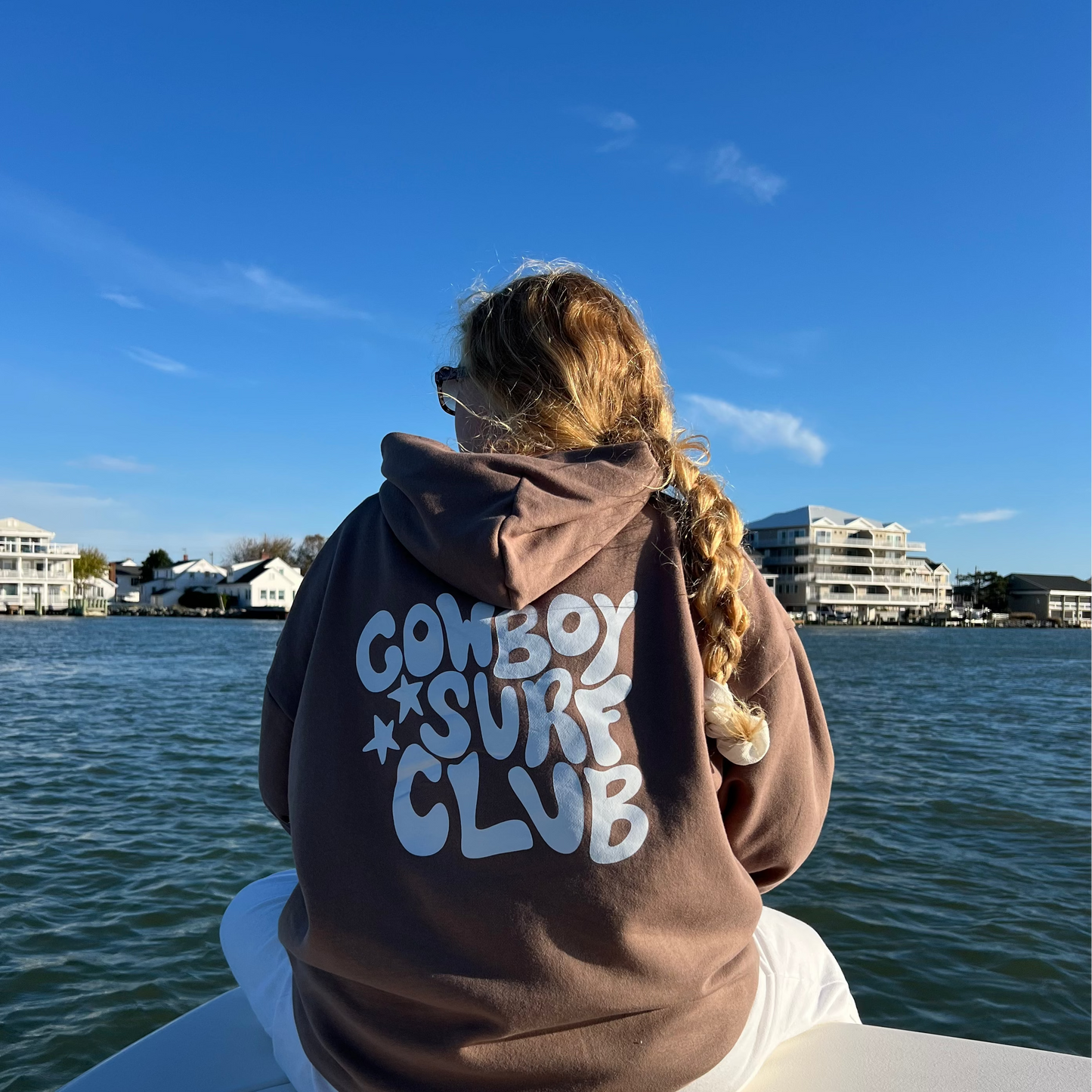 Person wearing a hoodie with 'Cowboy Surf Club' text on a boat by water
